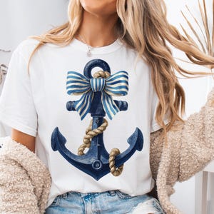 May include: White t-shirt featuring a navy blue anchor design with a striped bow and rope detailing. The anchor has a nautical theme. The design is centered on the front of the shirt.