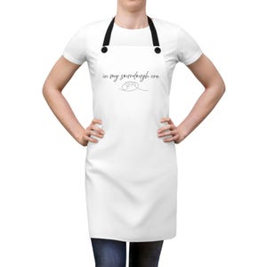 Pode incluir: Avental branco com alças pretas e o texto "in my sourdough era" em fonte cursiva. O avental tem um design simples, adequado para assar ou cozinhar. O avental é usado por uma pessoa.
