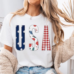 Pode incluir: Camiseta branca com a palavra "USA" em um design patriótico. A letra "U" é azul com estrelas brancas, "S" tem um padrão floral e "A" é xadrez vermelho e branco. A camiseta é usada por uma pessoa.