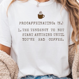 May include: White t-shirt with the text "PROCAFFEINATING: (N.) 1. THE TENDENCY TO NOT START ANYTHING UNTIL YOU'VE HAD COFFEE." A brown coffee cup graphic is above the text. The shirt is a casual style.
