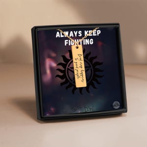 May include: A gold-colored rectangular pendant necklace with the words "Bad crap happens to good people" engraved on it. The necklace is displayed in a black box with the words "ALWAYS KEEP FIGHTING" on the background.