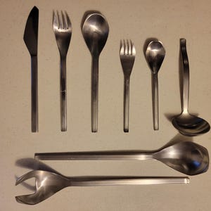 May include: A collection of stainless steel flatware, including a knife, forks, spoons, and a ladle. The utensils have a simple, modern design with long, slender handles. The arrangement includes serving spoons and a serving fork.