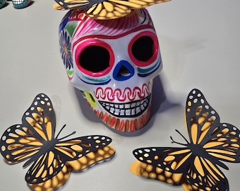 May include: A colorful, hand-painted ceramic skull with black butterfly decorations. The skull has pink, blue, yellow, and white paint with a whimsical design. The butterflies are layered and have a black and yellow color scheme.