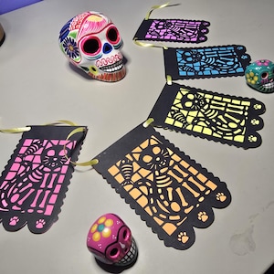 May include: A Day of the Dead themed paper banner with black paper flags and colorful cut-out skeletons of cats. The banner is strung with yellow ribbon. Two small sugar skull decorations are also included.