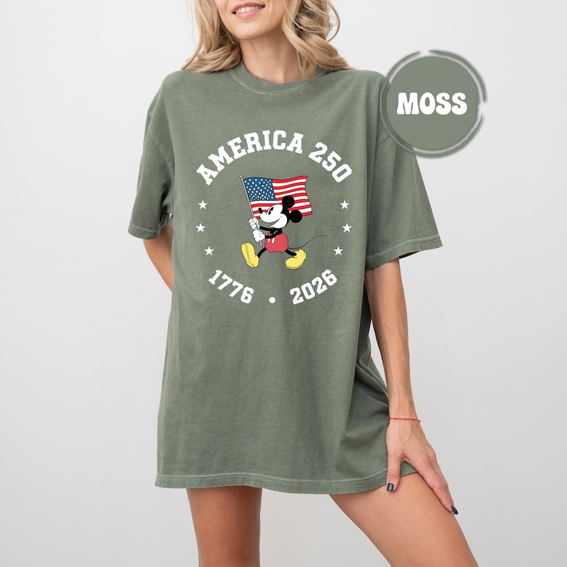 May include: Moss green t-shirt with a graphic of Mickey Mouse waving an American flag. The text "AMERICA 250" is arched above the character, with "1776" and "2026" below. The shirt has a loose fit.