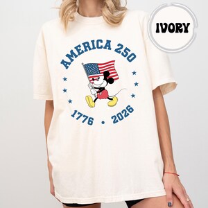 May include: Ivory-coloured t-shirt with a graphic of Mickey Mouse holding an American flag. The design includes the text "AMERICA 250" and the years "1776" and "2026", surrounded by stars. The word "IVORY" is in a circle at the top.