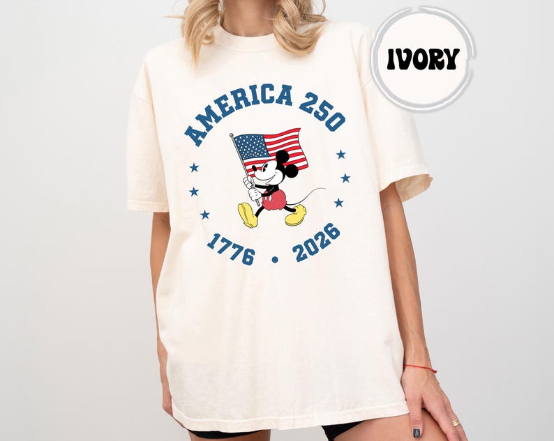 May include: Ivory-coloured t-shirt with a graphic of Mickey Mouse holding an American flag. The design includes the text "AMERICA 250" and the years "1776" and "2026", surrounded by stars. The word "IVORY" is in a circle at the top.