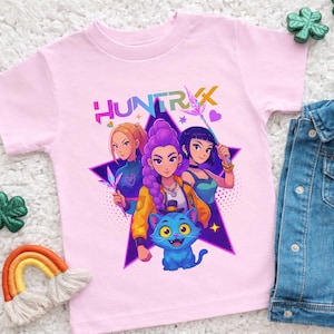 Peut inclure: T-shirt rose clair avec un motif de trois personnages féminins, un chat bleu et le mot "HUNTRIX" dans une police colorée. Les personnages sont placés sur une étoile violette. Une veste en jean et des objets décoratifs sont en arrière-plan.