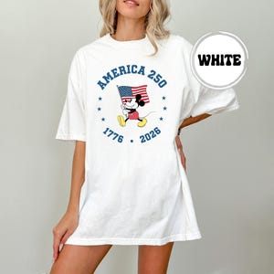 May include: White t-shirt with a graphic of Mickey Mouse waving an American flag. The design includes the text "America 250" and the years "1776" and "2026" in blue. The shirt has short sleeves and a loose fit.
