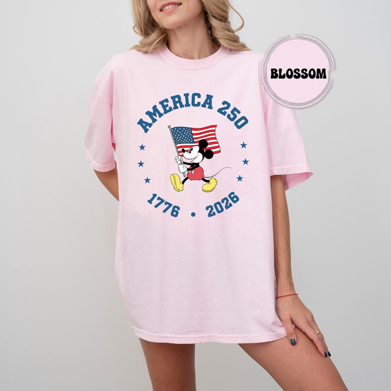 May include: A pale pink t-shirt with a cartoon Mickey Mouse design holding an American flag. The design includes the text "AMERICA 250" in blue, and the years "1776" and "2026".