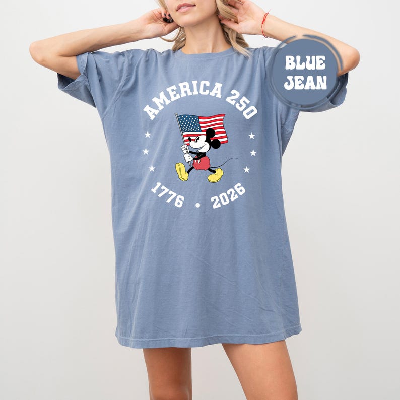May include: Blue jean-coloured oversized t-shirt with a graphic of Mickey Mouse holding an American flag. The text "AMERICA 250" arches above the character, with "1776" and "2026" below. White stars surround the design.