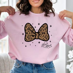 Peut inclure: Un sweat-shirt rose clair avec un motif de n&oelig;ud l&eacute;opard. Le n&oelig;ud est bord&eacute; de noir et entour&eacute; d'&eacute;toiles noires. Le texte manuscrit "Minnie Mouse xoxo" est imprim&eacute; sous le n&oelig;ud.