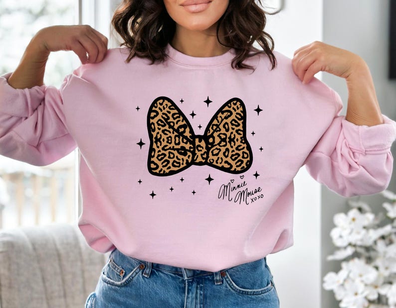 Peut inclure: Un sweat-shirt rose clair avec un motif de n&oelig;ud l&eacute;opard. Le n&oelig;ud est bord&eacute; de noir et entour&eacute; d'&eacute;toiles noires. Le texte manuscrit "Minnie Mouse xoxo" est imprim&eacute; sous le n&oelig;ud.