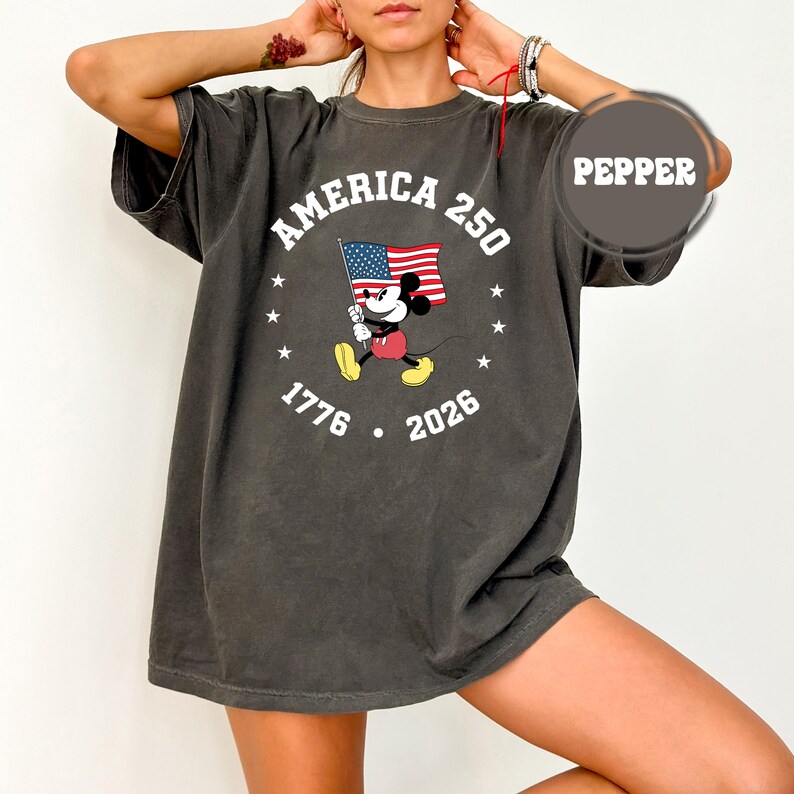 May include: Dark grey t-shirt with a graphic of Mickey Mouse holding an American flag. The design includes the text "AMERICA 250" and the years "1776" and "2026". The shirt has a loose fit and is a pepper color.