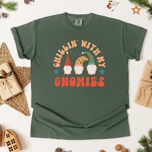 Puede incluir: Camiseta verde oscuro con la frase "Chillin' with my Gnomies" en naranja y blanco, adornada con tres gnomos. El dise&ntilde;o est&aacute; rodeado de decoraciones festivas, incluyendo una estrella, regalos y ramas de pino.