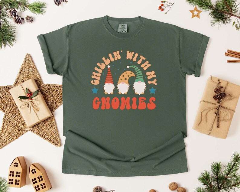 Puede incluir: Camiseta verde oscuro con la frase "Chillin' with my Gnomies" en naranja y blanco, adornada con tres gnomos. El dise&ntilde;o est&aacute; rodeado de decoraciones festivas, incluyendo una estrella, regalos y ramas de pino.