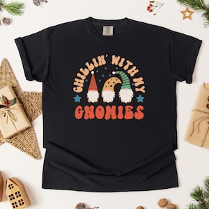 Puede incluir: Camiseta negra con el texto "CHILLIN' WITH MY GNOMIES" en naranja y blanco. El dise&ntilde;o presenta tres gnomos con sombreros coloridos. La camiseta est&aacute; rodeada de decoraciones navide&ntilde;as, incluyendo una estrella, regalos y ramas de pino.