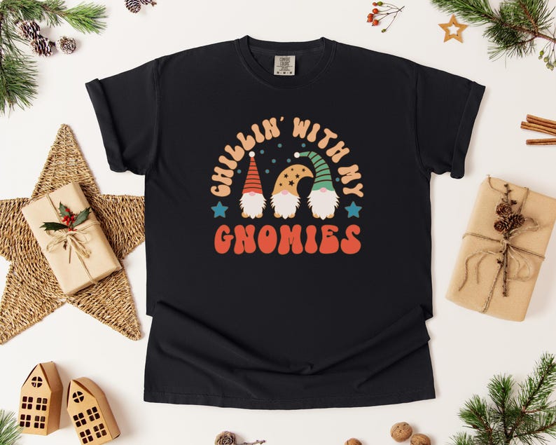 Puede incluir: Camiseta negra con el texto "CHILLIN' WITH MY GNOMIES" en naranja y blanco. El dise&ntilde;o presenta tres gnomos con sombreros coloridos. La camiseta est&aacute; rodeada de decoraciones navide&ntilde;as, incluyendo una estrella, regalos y ramas de pino.