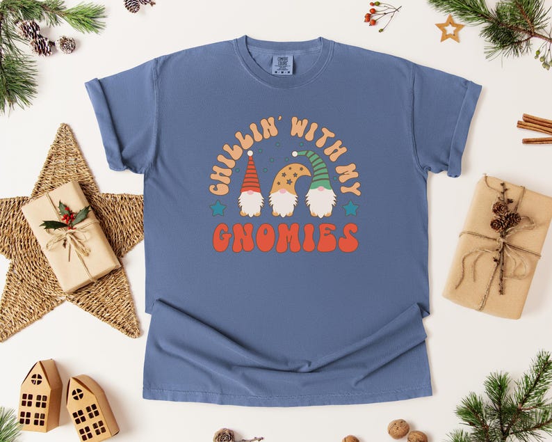 Puede incluir: Una camiseta azul con el texto "Chillin' with my Gnomies" en naranja, beige y verde. El gr&aacute;fico presenta tres gnomos con sombreros coloridos. La camiseta est&aacute; rodeada de decoraciones navide&ntilde;as.