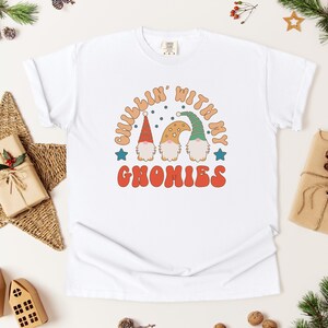 Puede incluir: Camiseta blanca con el texto "Chillin' with my Gnomies" en una fuente retro. El gr&aacute;fico presenta tres gnomos con sombreros coloridos. La camiseta est&aacute; rodeada de decoraciones navide&ntilde;as, incluyendo una estrella, regalos y vegetaci&oacute;n.