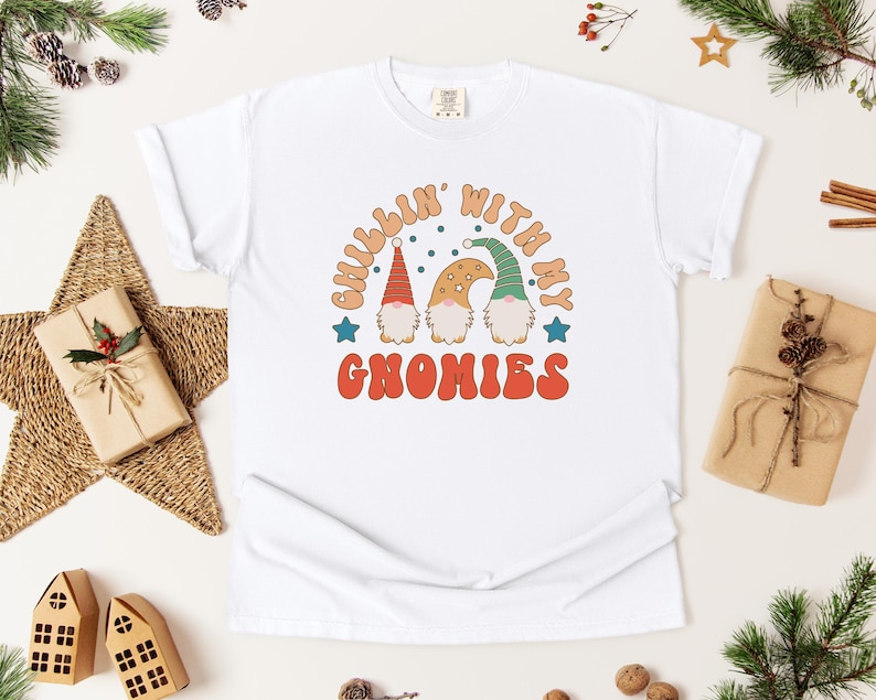 Puede incluir: Camiseta blanca con el texto "Chillin' with my Gnomies" en una fuente retro. El gr&aacute;fico presenta tres gnomos con sombreros coloridos. La camiseta est&aacute; rodeada de decoraciones navide&ntilde;as, incluyendo una estrella, regalos y vegetaci&oacute;n.