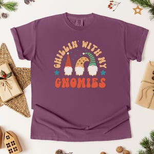 Puede incluir: Una camiseta morada con la frase "Chillin' with my Gnomies" en una fuente retro. El dise&ntilde;o presenta tres gnomos con sombreros coloridos. La camiseta est&aacute; rodeada de decoraciones navide&ntilde;as, incluyendo una estrella, regalos envueltos y ramas de pino.