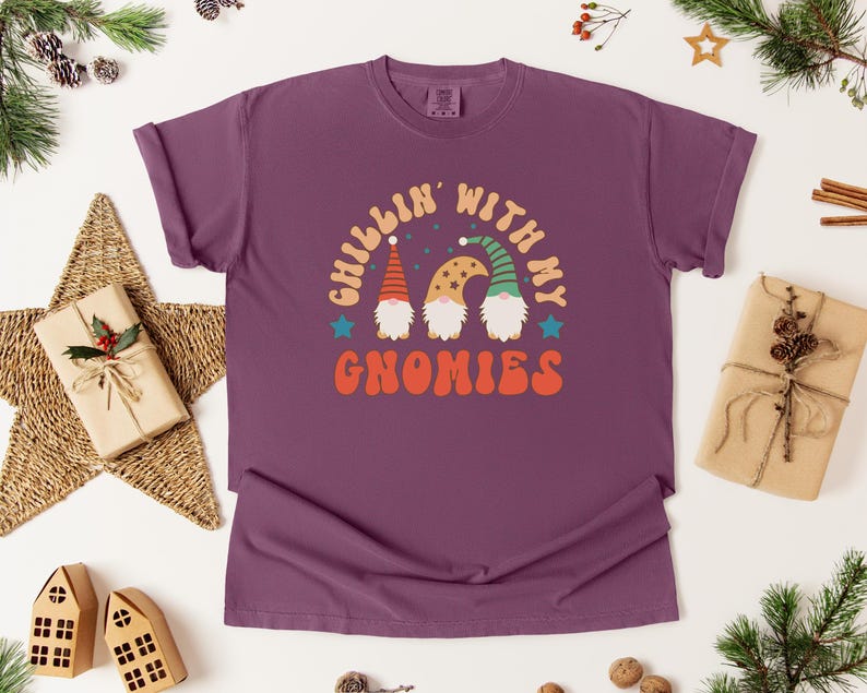 Puede incluir: Una camiseta morada con la frase "Chillin' with my Gnomies" en una fuente retro. El dise&ntilde;o presenta tres gnomos con sombreros coloridos. La camiseta est&aacute; rodeada de decoraciones navide&ntilde;as, incluyendo una estrella, regalos envueltos y ramas de pino.