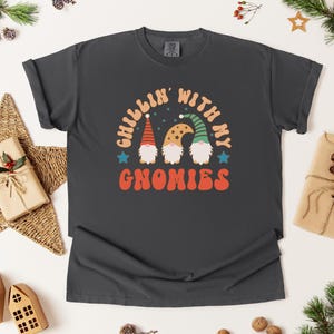 Puede incluir: Camiseta gris oscuro con el texto "Chillin' with my Gnomies" en naranja, blanco y azul. El gr&aacute;fico presenta tres gnomos con sombreros coloridos. La camiseta es de estilo c&oacute;modo e informal, perfecta para la temporada navide&ntilde;a.