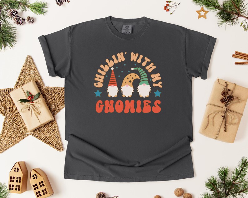 Puede incluir: Camiseta gris oscuro con el texto "Chillin' with my Gnomies" en naranja, blanco y azul. El gr&aacute;fico presenta tres gnomos con sombreros coloridos. La camiseta es de estilo c&oacute;modo e informal, perfecta para la temporada navide&ntilde;a.