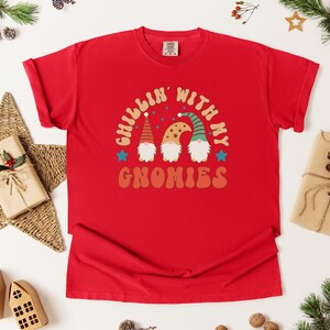 Puede incluir: Camiseta roja con la frase "Chillin' with my Gnomies". El dise&ntilde;o presenta tres gnomos con sombreros y barbas coloridas. La camiseta est&aacute; rodeada de decoraciones navide&ntilde;as.