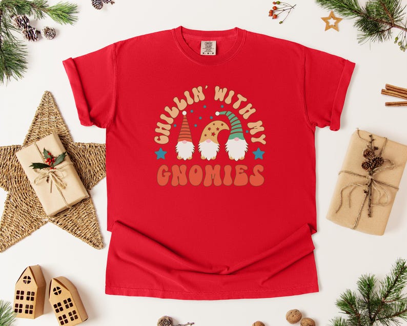 Puede incluir: Camiseta roja con la frase "Chillin' with my Gnomies". El dise&ntilde;o presenta tres gnomos con sombreros y barbas coloridas. La camiseta est&aacute; rodeada de decoraciones navide&ntilde;as.