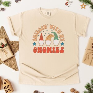 Puede incluir: Camiseta color crema con el texto "Chillin' with my Gnomies" en una fuente retro. El gr&aacute;fico presenta tres gnomos con sombreros rojos, beige y verdes. La camiseta es de estilo casual y unisex.
