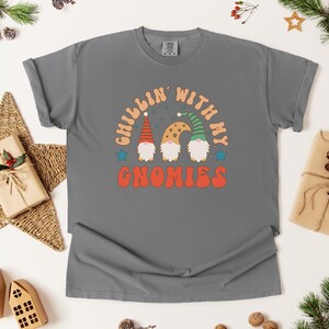 Puede incluir: Una camiseta gris con el texto "Chillin' with my Gnomies" en una fuente retro. El gr&aacute;fico presenta tres gnomos con sombreros coloridos. La camiseta est&aacute; rodeada de decoraciones navide&ntilde;as, incluyendo regalos envueltos y ramas de pino.