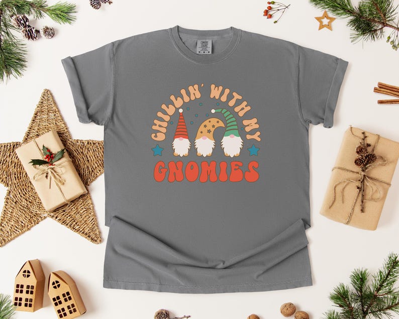 Puede incluir: Una camiseta gris con el texto "Chillin' with my Gnomies" en una fuente retro. El gr&aacute;fico presenta tres gnomos con sombreros coloridos. La camiseta est&aacute; rodeada de decoraciones navide&ntilde;as, incluyendo regalos envueltos y ramas de pino.