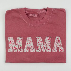 May include: A pink t-shirt with the word "MAMA" spelled out in large white letters. The letters are covered in a floral pattern.
