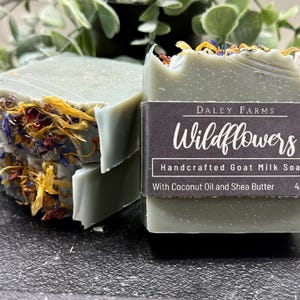 May include: Stacked bars of handcrafted goat milk soap with dried wildflowers. The soap is a pale green color and is decorated with colorful flower petals. The label reads "Wildflowers" and "4.5oz".