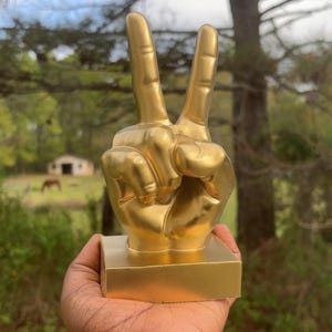 May include: A gold-colored peace sign hand sculpture on a gold-colored base. The sculpture is being held in a person's hand.
