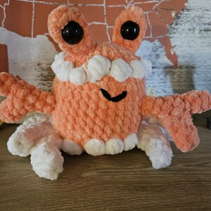 May include: A handmade, plush crab toy in shades of peach and white. The crab features large, black, round eyes, a row of white teeth, and multiple arms and legs. The toy is made of soft, textured yarn.