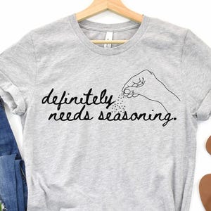 May include: A light gray t-shirt with the text "definitely needs seasoning" in black script. The design includes a hand sprinkling seasoning. The shirt is displayed with blue jeans, white flowers, and brown sandals.