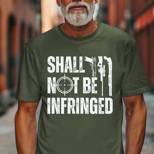 May include: Military green t-shirt with the text "SHALL NOT BE INFRINGED" in white. The design includes a crosshair, a handgun, and a rifle. The shirt is a crew neck style.