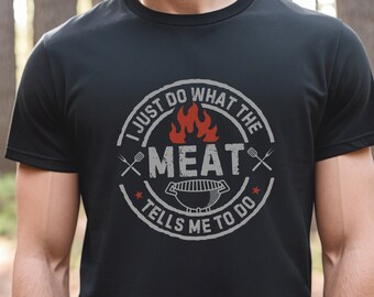May include: Black t-shirt with a white circle graphic that says "I just do what the meat tells me to do". The graphic also includes a grill, flames, and two crossed grilling tools.