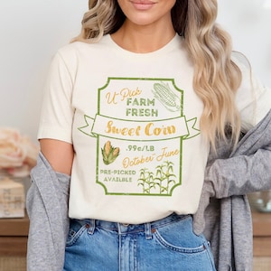 May include: A white t-shirt with a vintage-style graphic of a corn cob and the text "U-Pick Farm Fresh Sweet Corn .99c/LB October June Pre-Picked Available".