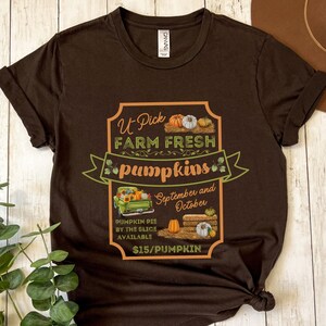 May include: A brown t-shirt with a graphic of a sign that says "U Pick Farm Fresh Pumpkins". The sign is decorated with pumpkins, hay bales, and a truck. The text on the sign also says "September and October" and "$15/Pumpkin".