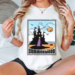 Pu&ograve; includere: Maglietta bianca con una grafica di un castello nero su una spiaggia con una cascata viola e il testo "Summerween".