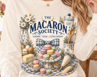 Peut inclure: Un t-shirt à manches longues crème avec une impression graphique The Macaron Society, des motifs floraux bleus et blancs, une pile de macarons colorés, un bloc de beurre, un fouet, une poche à douille et le texte Almonds Sugar A Little Patience.