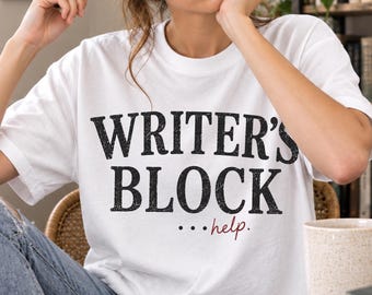 May include: A white short-sleeved t-shirt featuring the black distressed text WRITER'S BLOCK and the word help in a smaller red script font. The shirt has a crew neckline and a standard fit, providing a clean look for casual daily wear.