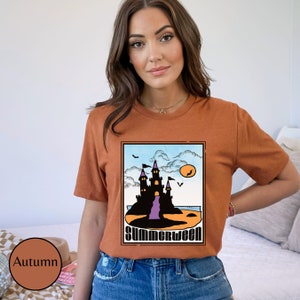 Pu&ograve; includere: Una maglietta color ruggine con una grafica di un castello nero su una spiaggia con un tramonto viola e arancione. Il testo "Summerween" &egrave; stampato sotto il castello.