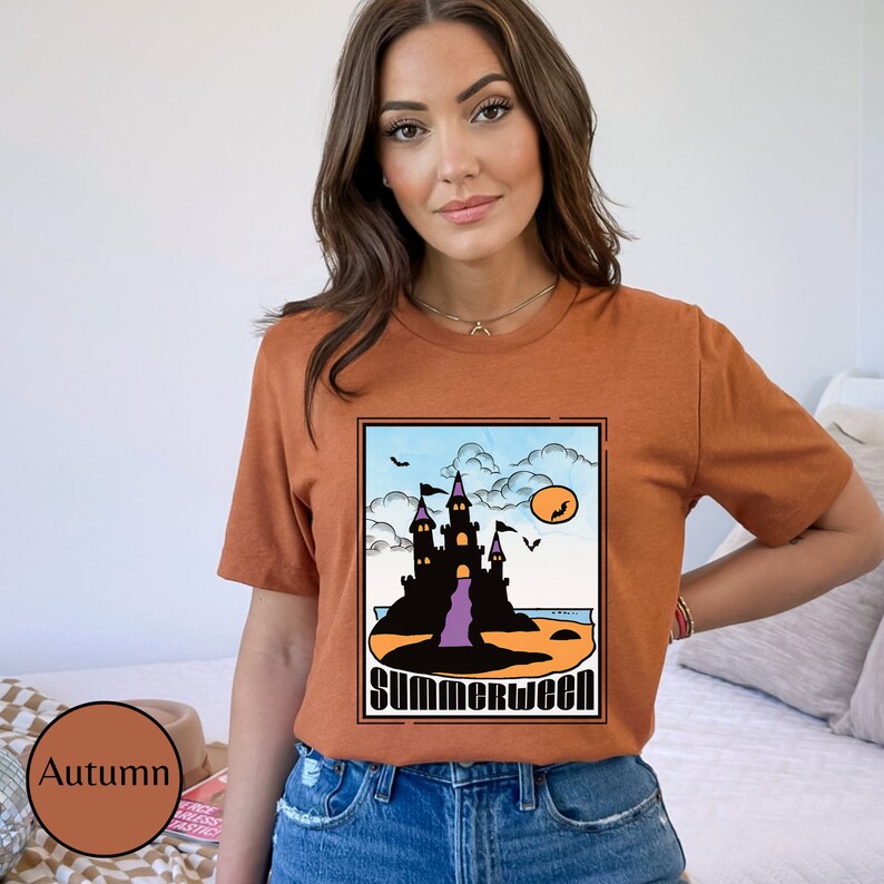 Pu&ograve; includere: Una maglietta color ruggine con una grafica di un castello nero su una spiaggia con un tramonto viola e arancione. Il testo "Summerween" &egrave; stampato sotto il castello.