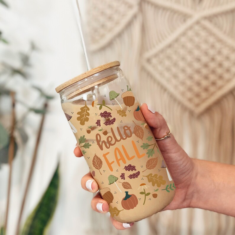 May include: A glass tumbler with a bamboo lid and a straw. The tumbler has a fall-themed design with leaves, acorns, and pumpkins. The text "hello fall" is printed on the tumbler.
