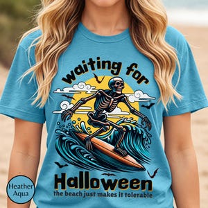 May include: Heather aqua t-shirt with a Halloween theme. The shirt features a skeleton surfing on a wave with the words "waiting for Halloween" and "the beach just makes it tolerable".